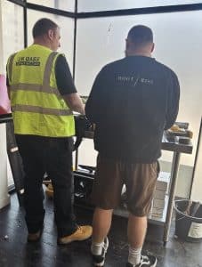 Two workers wearing high-visibility jackets and helmets standing next to a white industrial van on a construction site with scaffolding and building materials in the background.