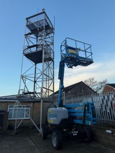 Blue Genie aerial work platform parked next to a tall metal scaffold tower under a clear sky